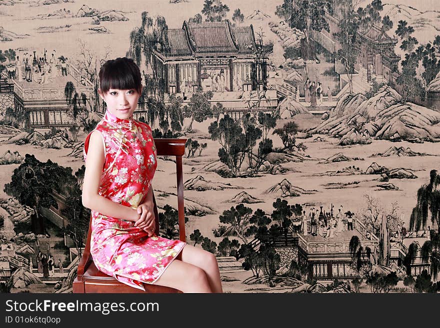 A Chinese girl in traditional dress sits in front of a Chinese old painting.