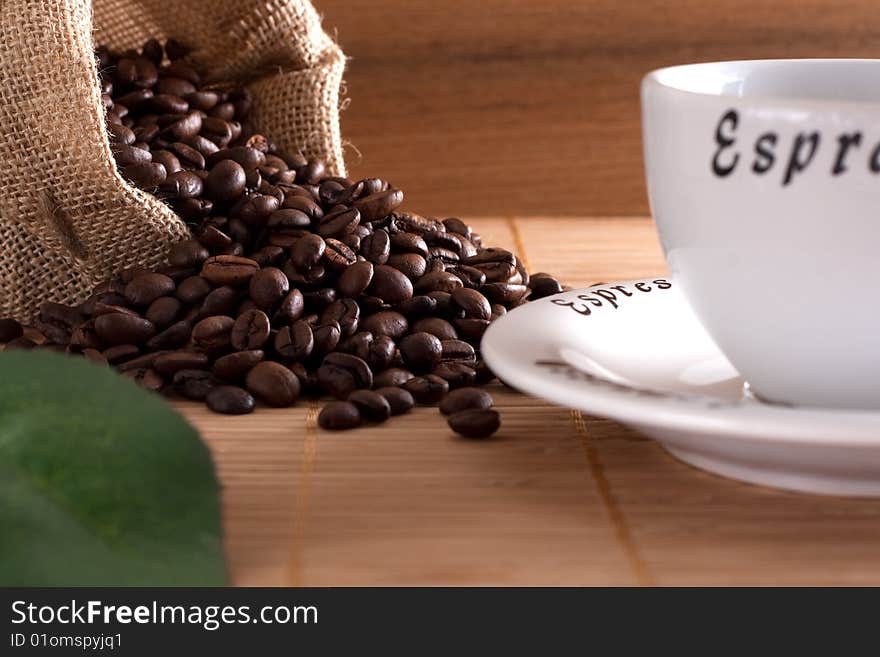 A bunch of coffee beans, falling out of a sack and a cup of fresh coffee on a wooden background