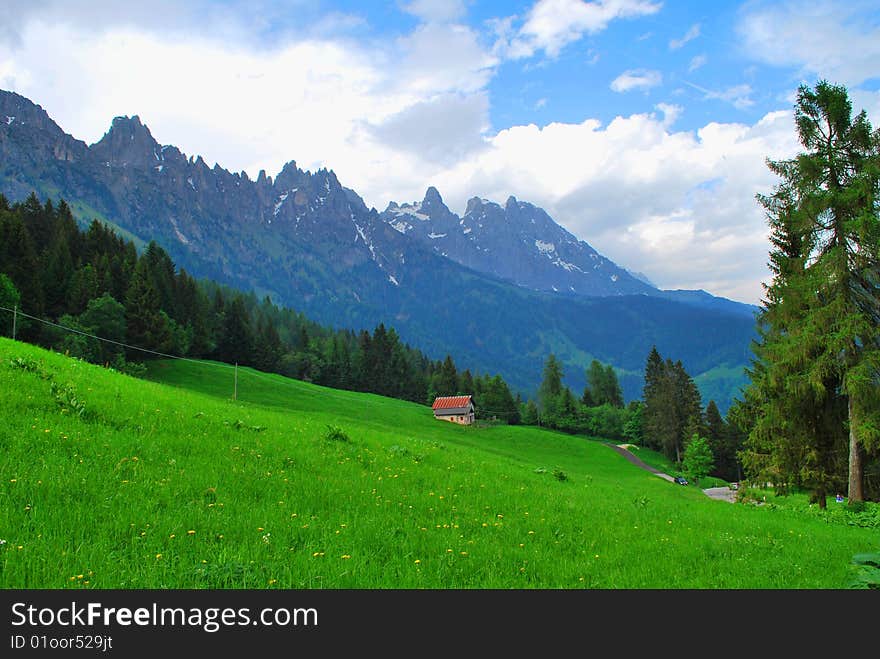 Plateau in the Alpes. Spring. A high-mountainous meadow on a background of rocky tops. The meadow is framed by a wood. On a meadow – country  construction. Plateau in the Alpes. Spring. A high-mountainous meadow on a background of rocky tops. The meadow is framed by a wood. On a meadow – country  construction.