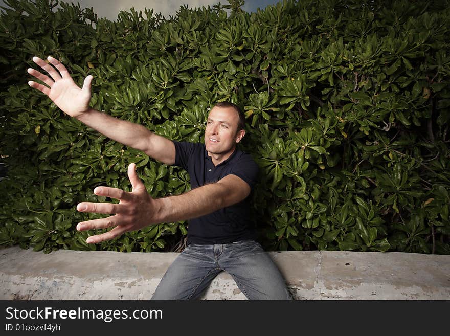 Young man sitting on a ledge by bushes and reaching out his arms