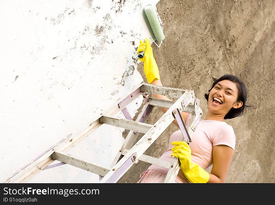 Cheerful asian young woman standing on a ladder, painting exterior wall of her house with a white colored paint, using a roller and gloves on hands. Cheerful asian young woman standing on a ladder, painting exterior wall of her house with a white colored paint, using a roller and gloves on hands