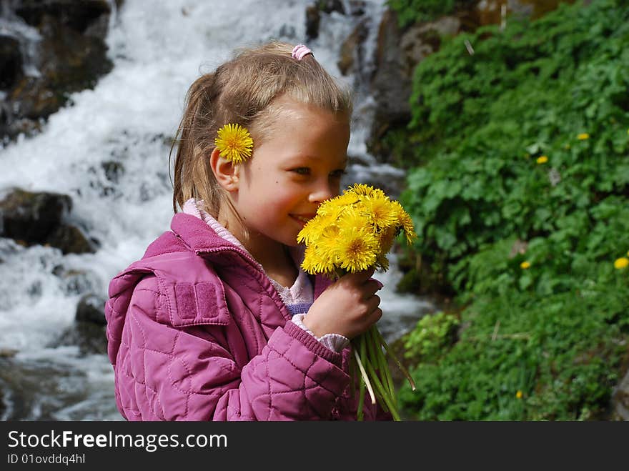 A little girl smelling yellow flowers and smiling, with a pink coat. In the background there is waterfall and green plants. A little girl smelling yellow flowers and smiling, with a pink coat. In the background there is waterfall and green plants.