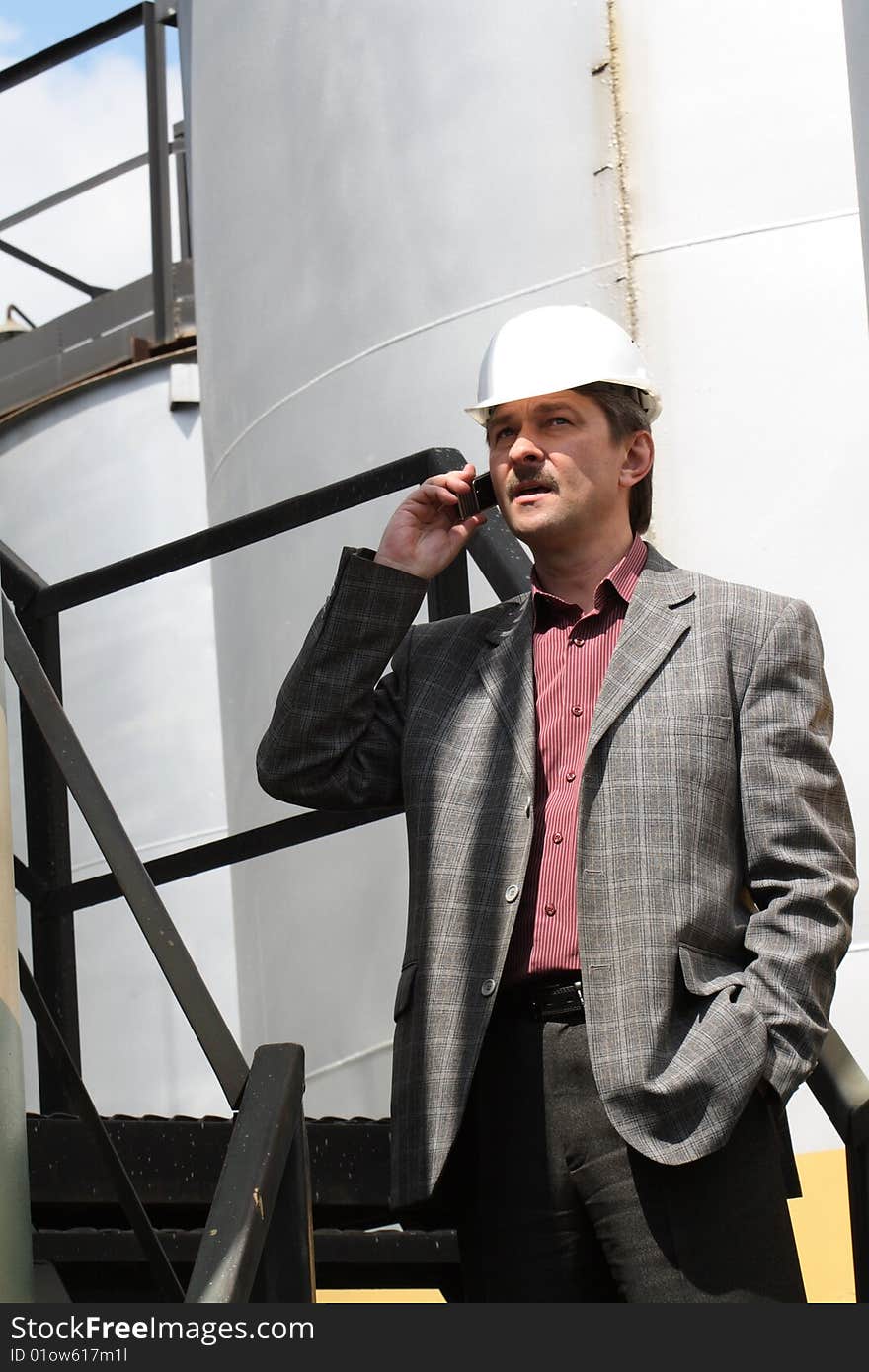 Engineer talking in phone, wearing hard-hat, on background with storage tank. Engineer talking in phone, wearing hard-hat, on background with storage tank