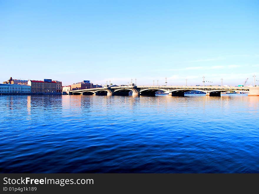 Photo of the city bridge through the magic dark blue-blue river against the equal sky. Photo of the city bridge through the magic dark blue-blue river against the equal sky