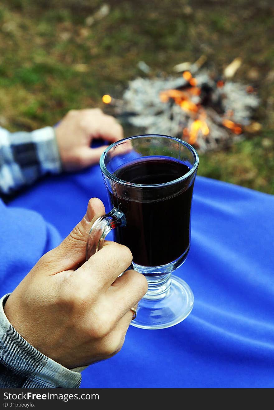 A mug with hot mulled wine in a hand of a man sitting near camp-fire under blue blanket at a picnic, blur background. A mug with hot mulled wine in a hand of a man sitting near camp-fire under blue blanket at a picnic, blur background