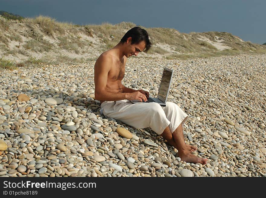 A young man sited on the beach working with a laptop. A young man sited on the beach working with a laptop