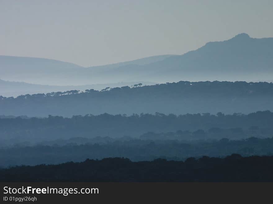 Early morning day break at the Huluhluwe-Umfolozi Game Reserve, South Africa, showing misty hills and silhouettes of trees. Early morning day break at the Huluhluwe-Umfolozi Game Reserve, South Africa, showing misty hills and silhouettes of trees