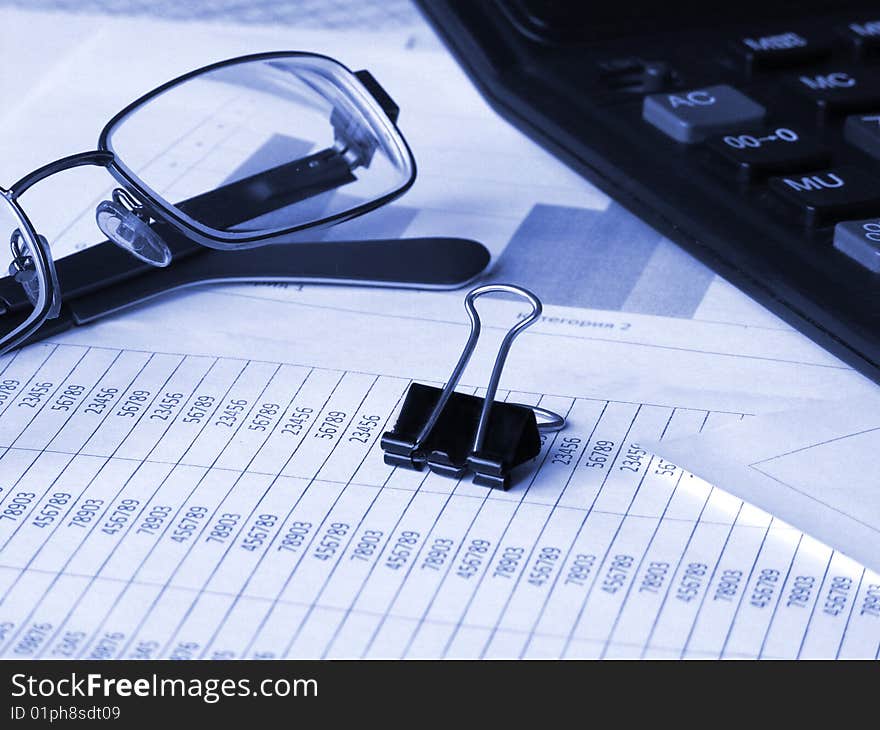 Glasses, calculator and binder clip on financial documents. Toned blue. Shallow DOF.