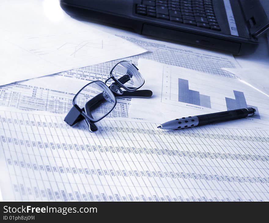 Laptop, glasses and pen on financial documents. Toned blue. Shallow DOF.