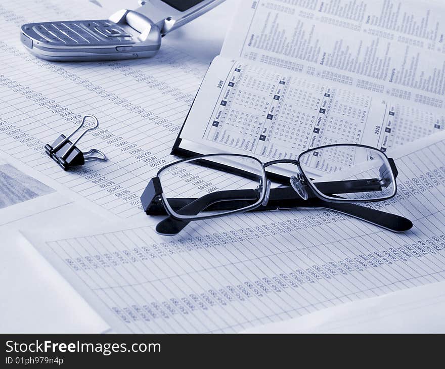 Glasses, daily book and mobile phone on financial documents. Toned blue. Shallow DOF.