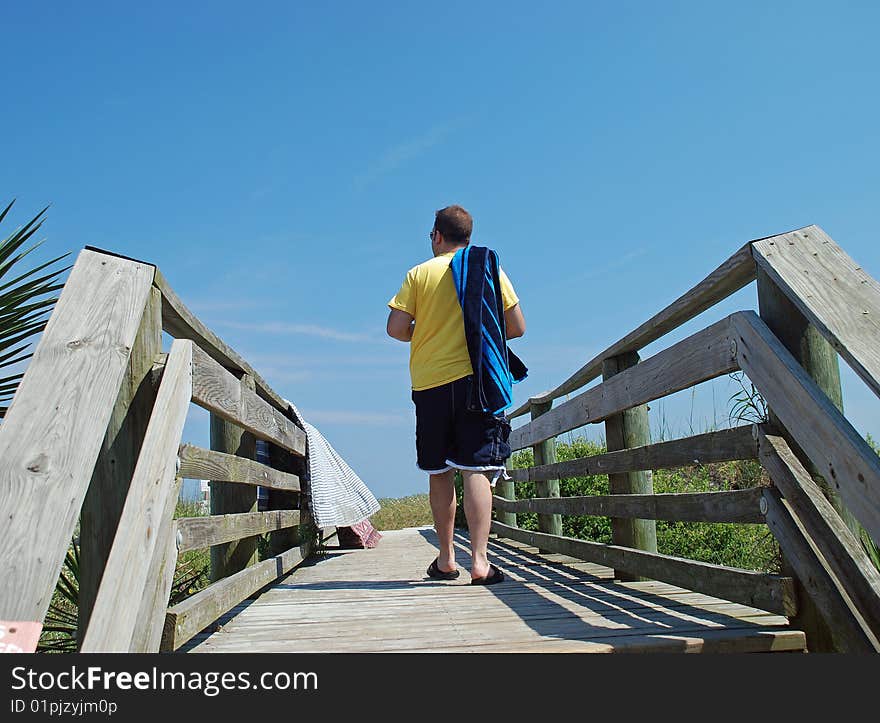 A vacationing man, carrying his towel over his shoulder, is walking along a boardwalk on his way to the beach. A vacationing man, carrying his towel over his shoulder, is walking along a boardwalk on his way to the beach.