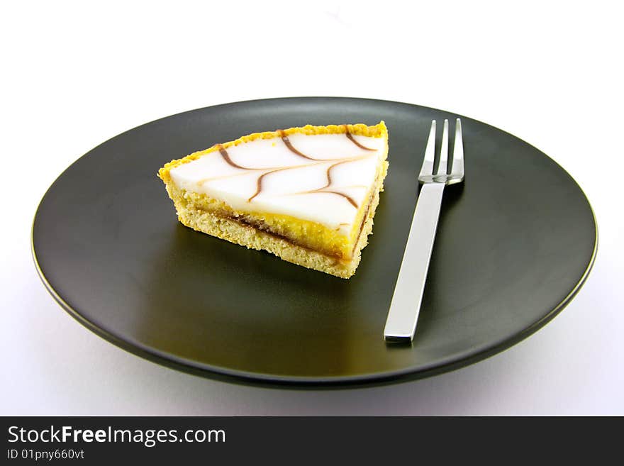 Delicious looking iced bakewell tart on a black plate with a fork and a plain background