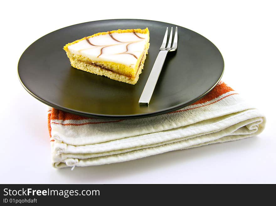 Delicious looking iced bakewell tart on a black plate with a fork and a plain background