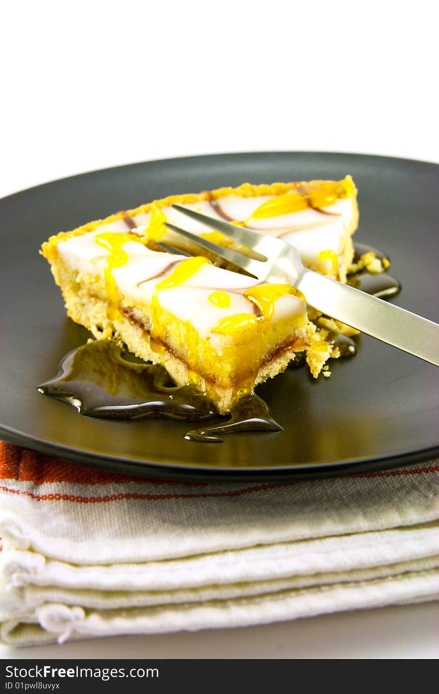 Delicious looking iced bakewell tart on a black plate with a treacle drizzle and a fork on a plain background