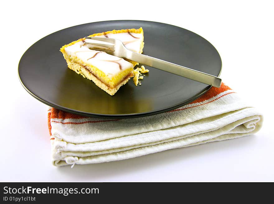 Delicious looking iced bakewell tart on a black plate with a fork and a plain background