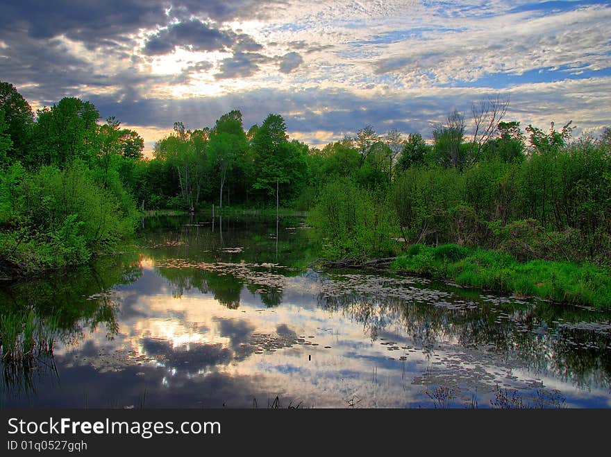 Clouds are reflected on the surface of a still pond as the sun is starting to shine through. Clouds are reflected on the surface of a still pond as the sun is starting to shine through.