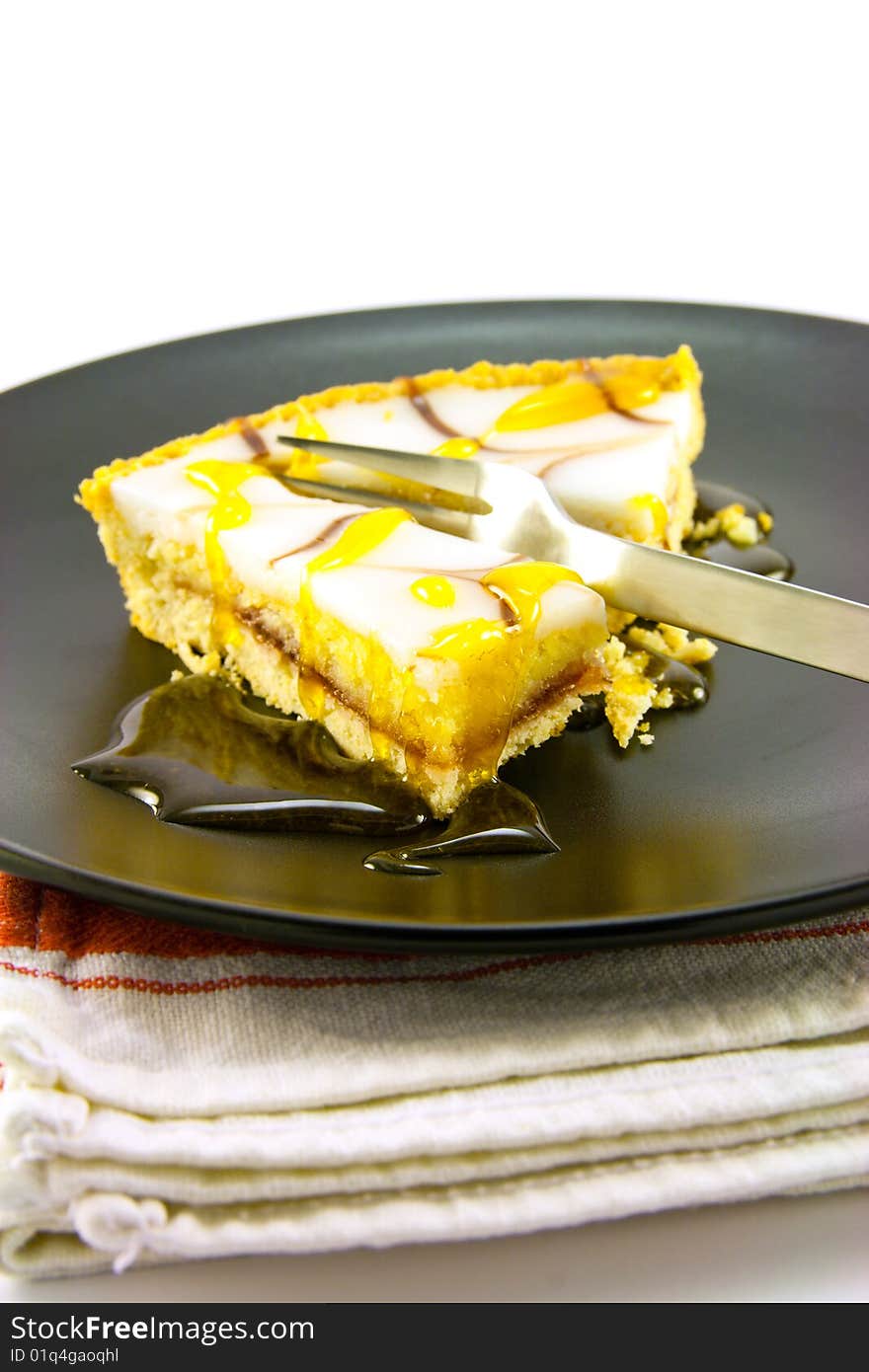 Delicious looking iced bakewell tart on a black plate with a treacle drizzle and a fork on a plain background