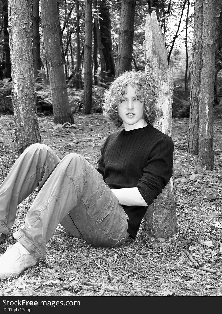 A black and white image of a teen sitting beside a tree in the woods. A black and white image of a teen sitting beside a tree in the woods