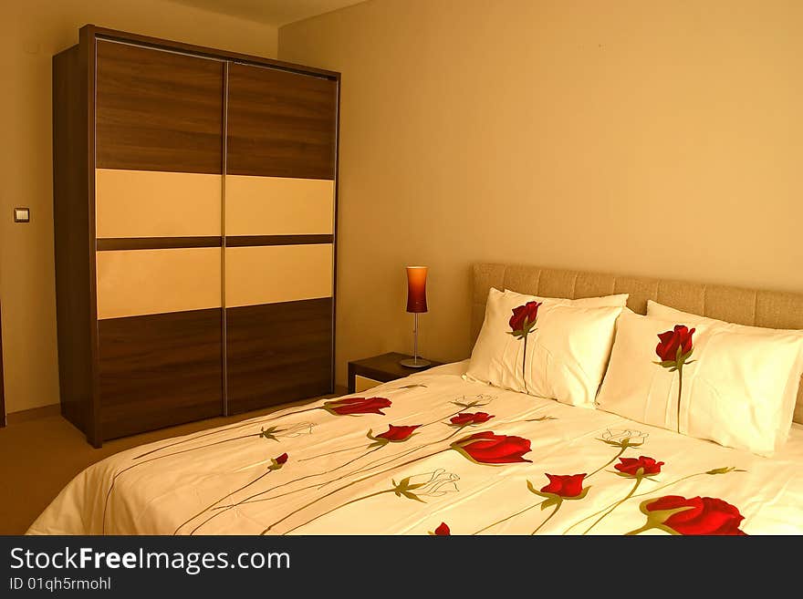 Warm bedroom with wardrobe and large bed with pillows and roses sheets. Warm bedroom with wardrobe and large bed with pillows and roses sheets.