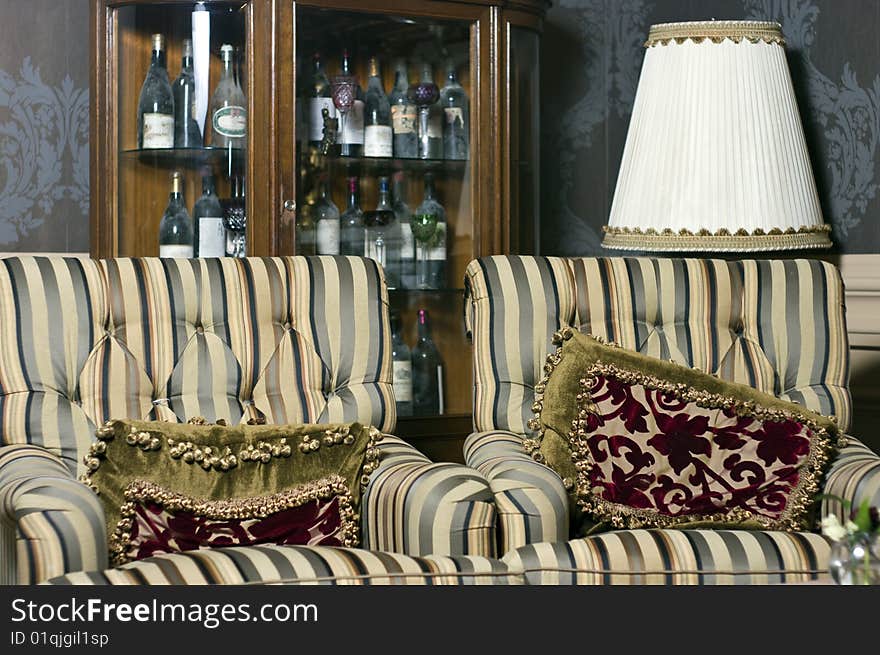 Interior of a retro, vintage room with two armchairs with cushions and cabinet with wine bottles behind. Interior of a retro, vintage room with two armchairs with cushions and cabinet with wine bottles behind.