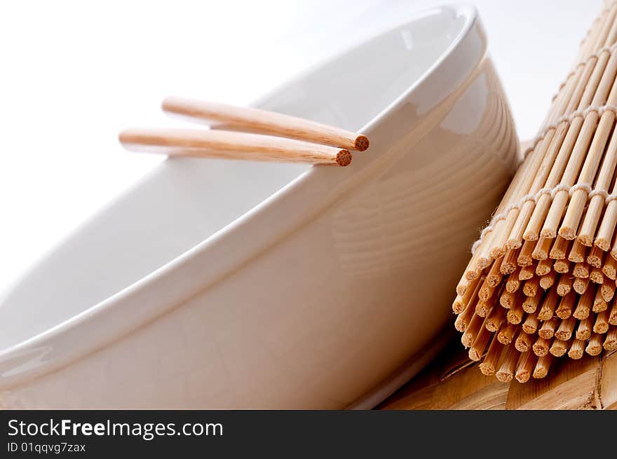 A tilted horizontal image of a pair of chopsticks on a white bowl with a sushi rolling mat on a weaved place mat