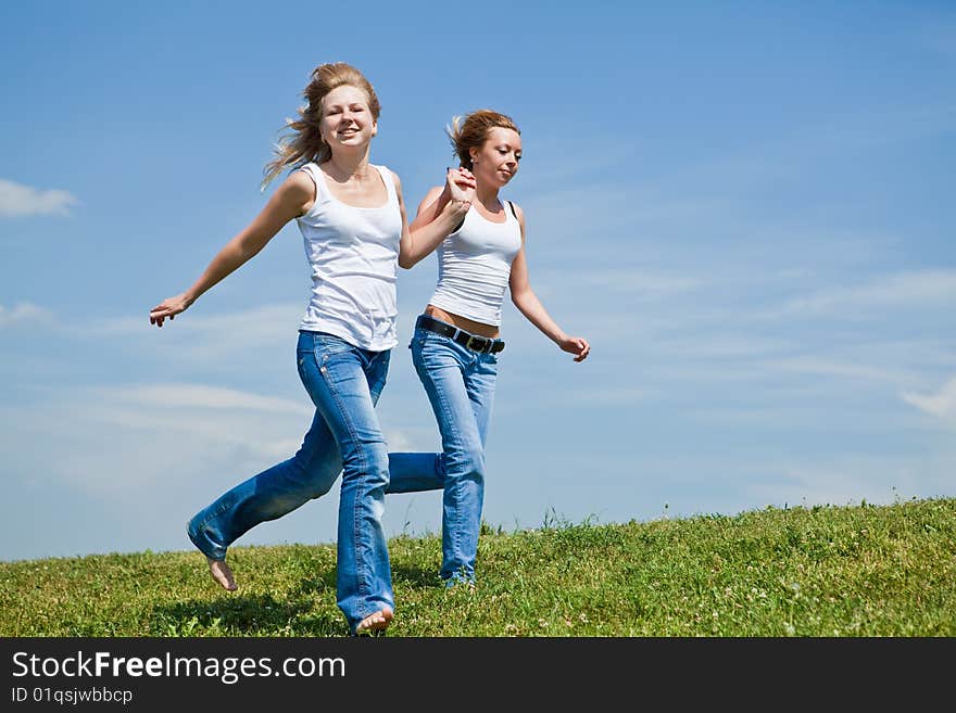 Two girls run on a green grass against the blue sky