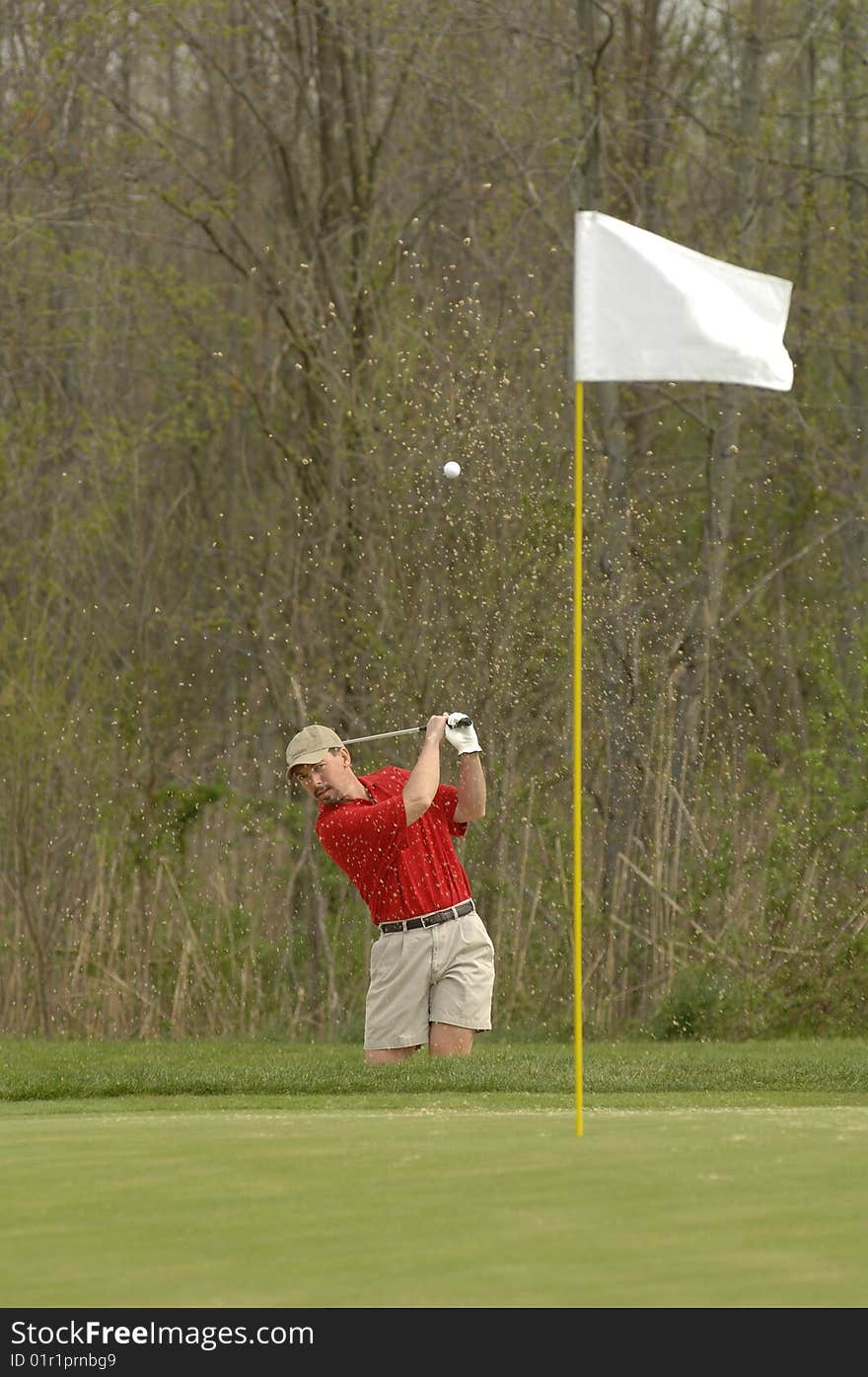 A man blasts out of a bunker kicking up sand. A man blasts out of a bunker kicking up sand