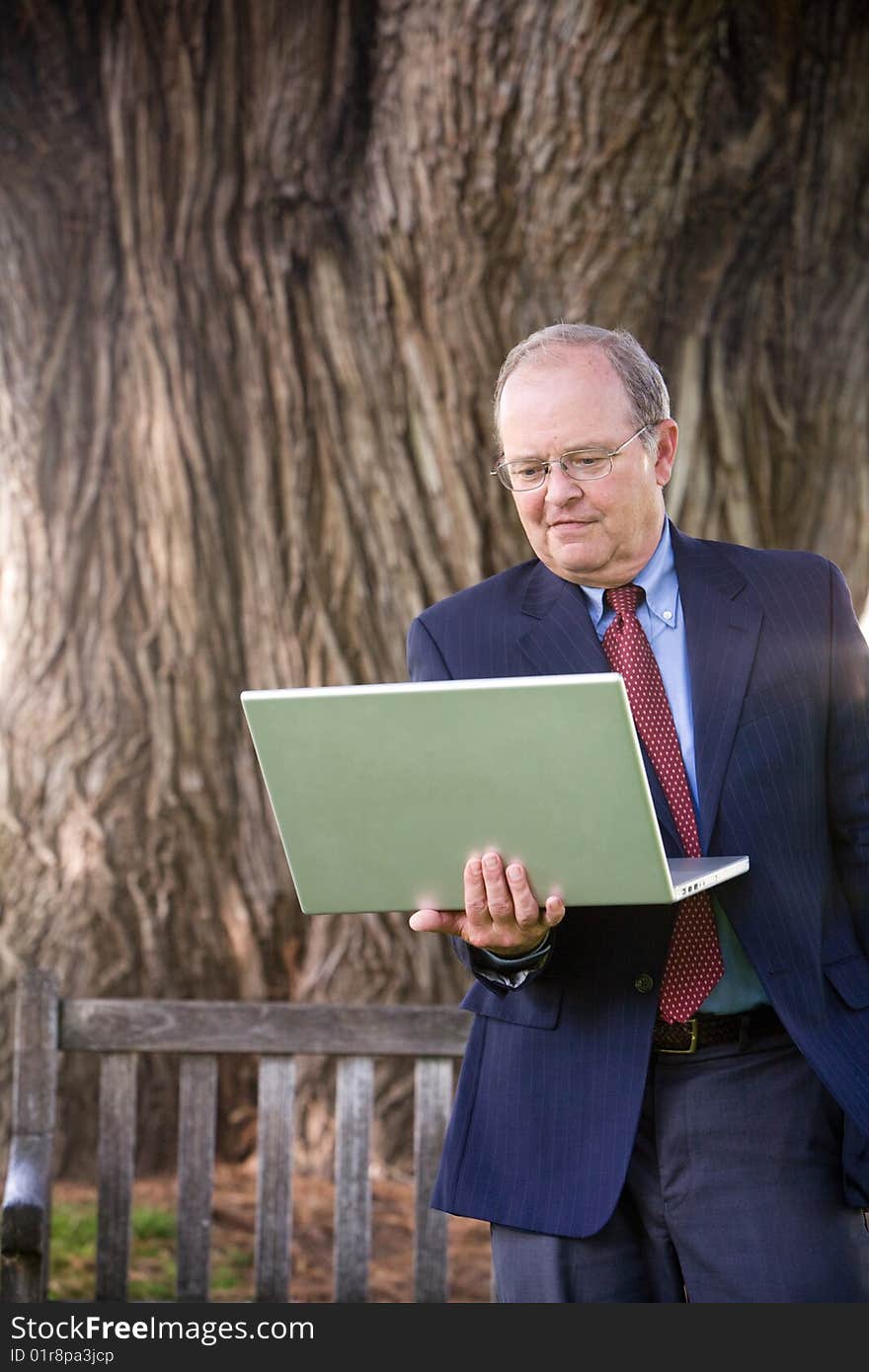 An older business man stands in the park while working on his laptop computer. An older business man stands in the park while working on his laptop computer