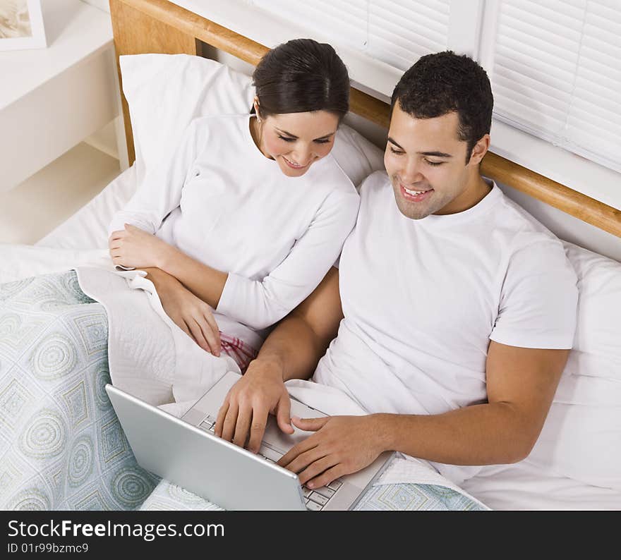 An attractive young couple lying in bed together and viewing a laptop screen.  They are both smiling. Horizontally framed shot. An attractive young couple lying in bed together and viewing a laptop screen.  They are both smiling. Horizontally framed shot.