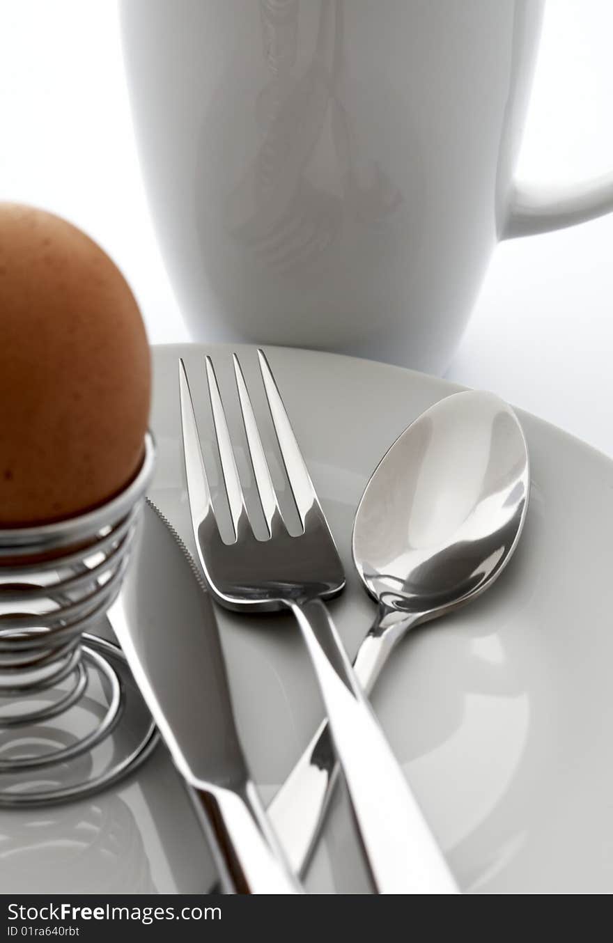 Egg, knife, fork, spoon on a white plate against a white cup. Egg, knife, fork, spoon on a white plate against a white cup