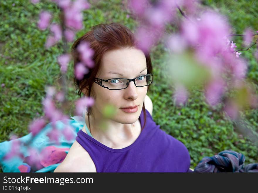 Young woman with glasses in park sitting on grass. Blured flowers in front. Young woman with glasses in park sitting on grass. Blured flowers in front.