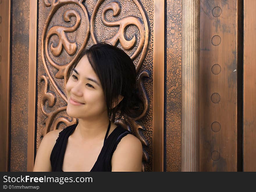 A young Chinese woman leaning on an old gloden cuprum door with a sweet smile.