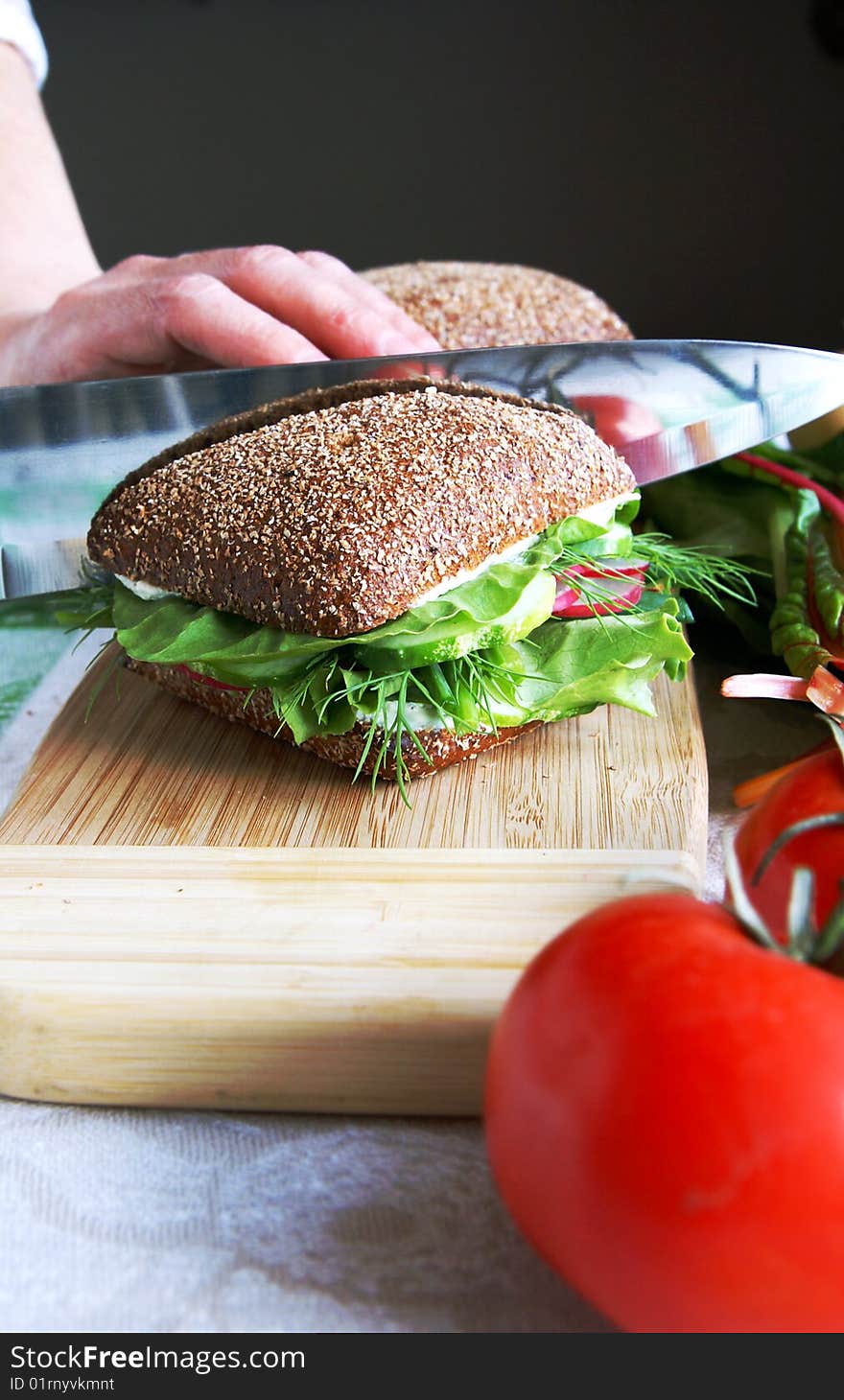 Housewife divides healthy rye bread sandwich with fresh radish, cucumber, green vegetables and herbs in half on a wooden kitchen board, blur black background, vertical. Housewife divides healthy rye bread sandwich with fresh radish, cucumber, green vegetables and herbs in half on a wooden kitchen board, blur black background, vertical