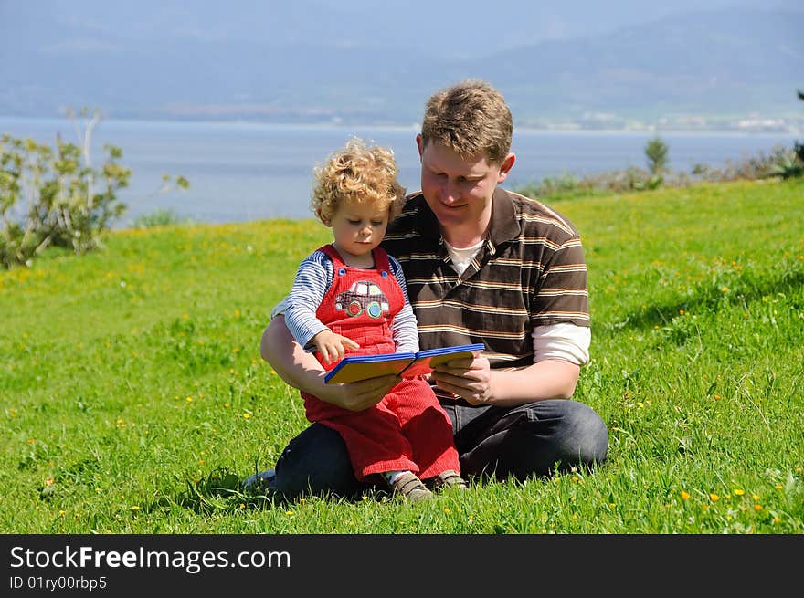 Father and son reading a book sitting on a green grass