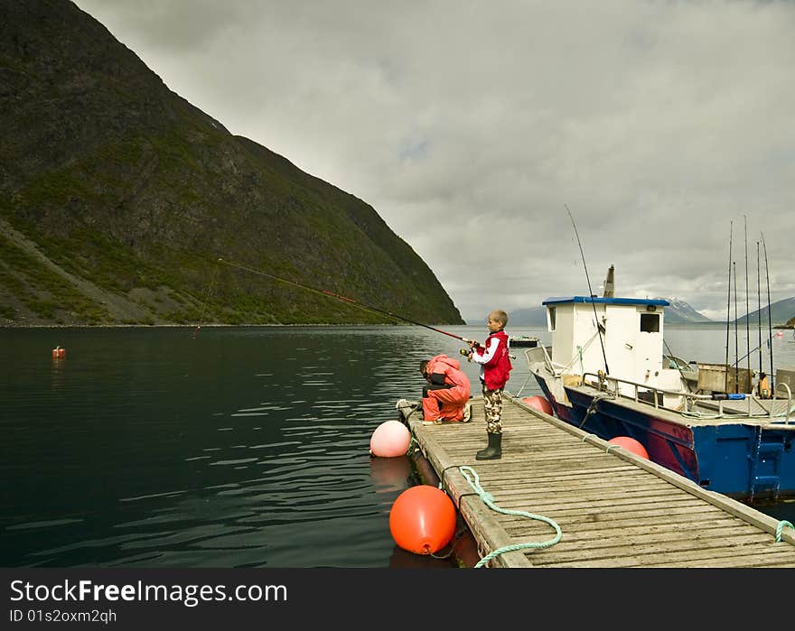 Two boys fishing in a fjord, standing on a wooden jetty next to a boat.