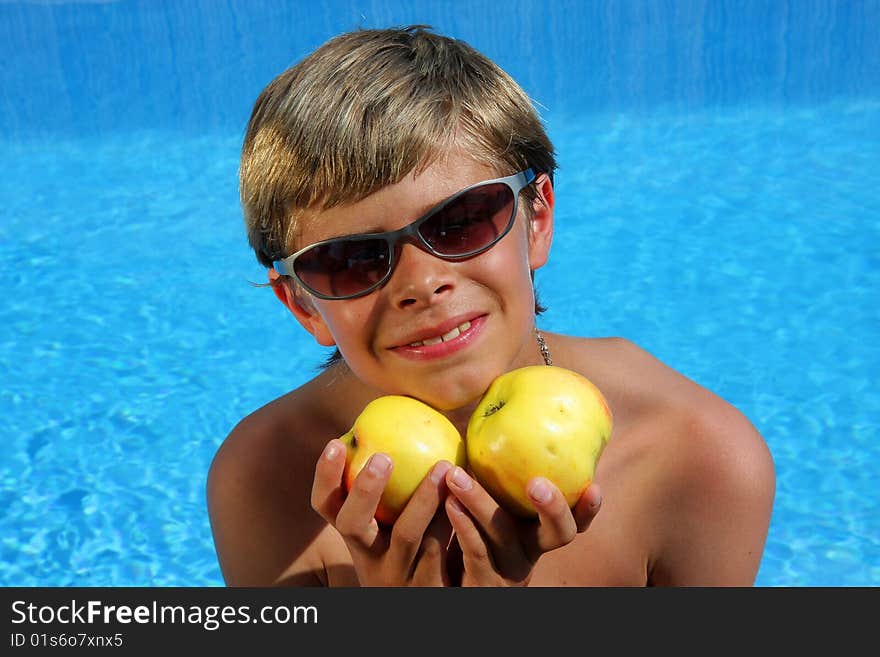 A 10 year old smiling American - German boy with sun glasses sitting at a swimming pool and presenting delicious apples in the summer sun. A 10 year old smiling American - German boy with sun glasses sitting at a swimming pool and presenting delicious apples in the summer sun