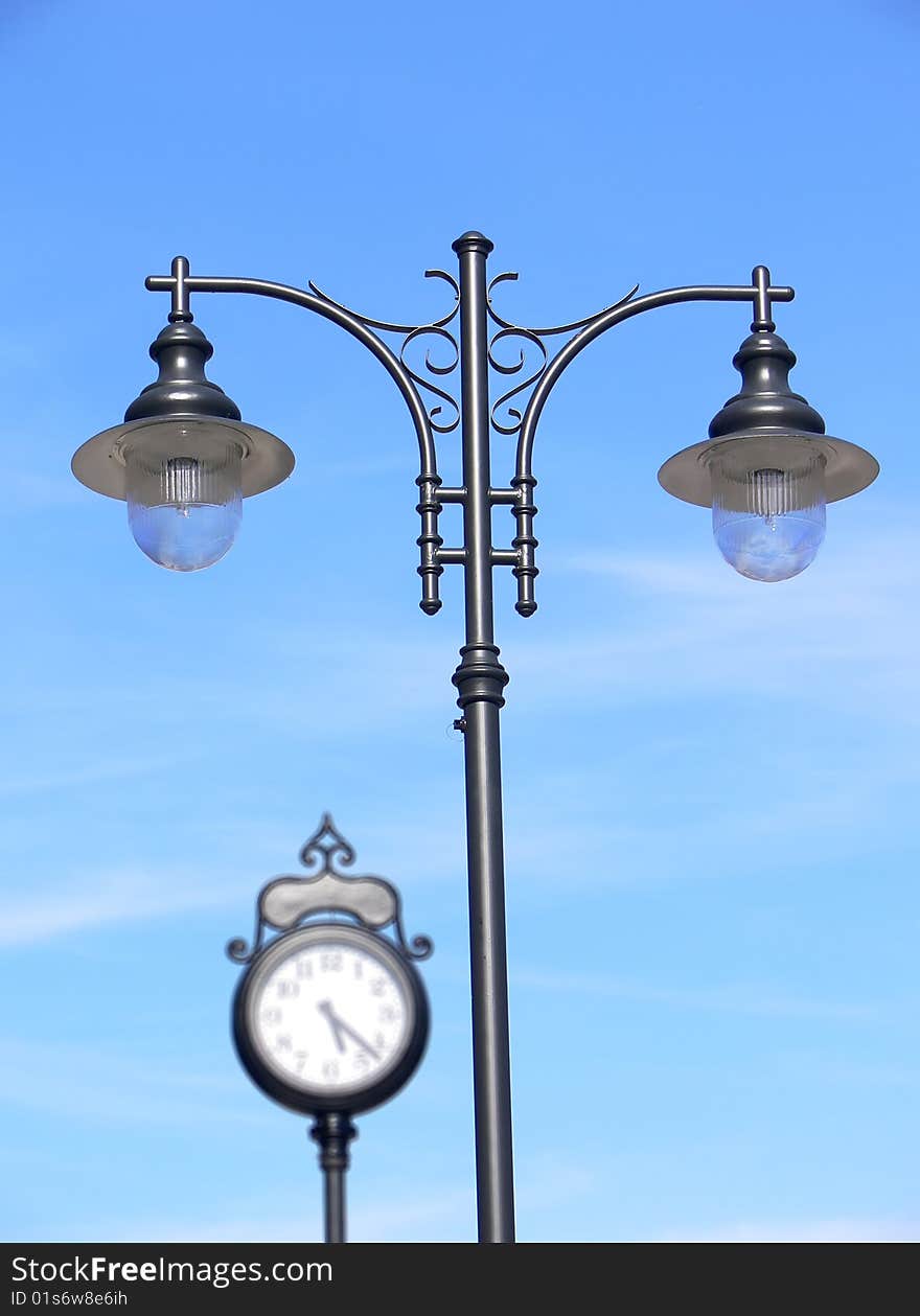 Closeup of the retro lamppost on the blue sky with blurred classic urban clock in the background. Closeup of the retro lamppost on the blue sky with blurred classic urban clock in the background.