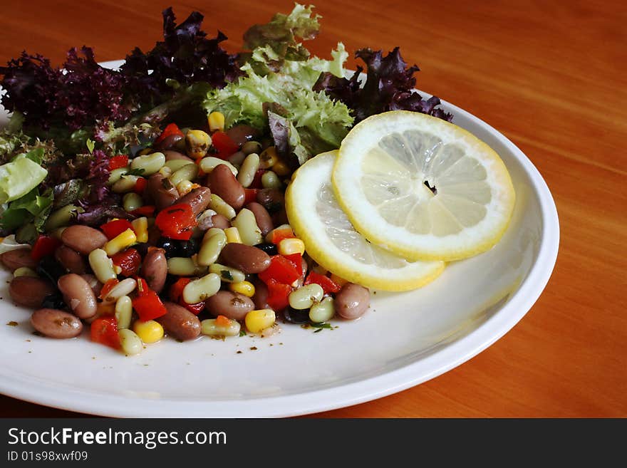 A white plate of mixed bean and lettuce salad with slices of lemon on a wood table