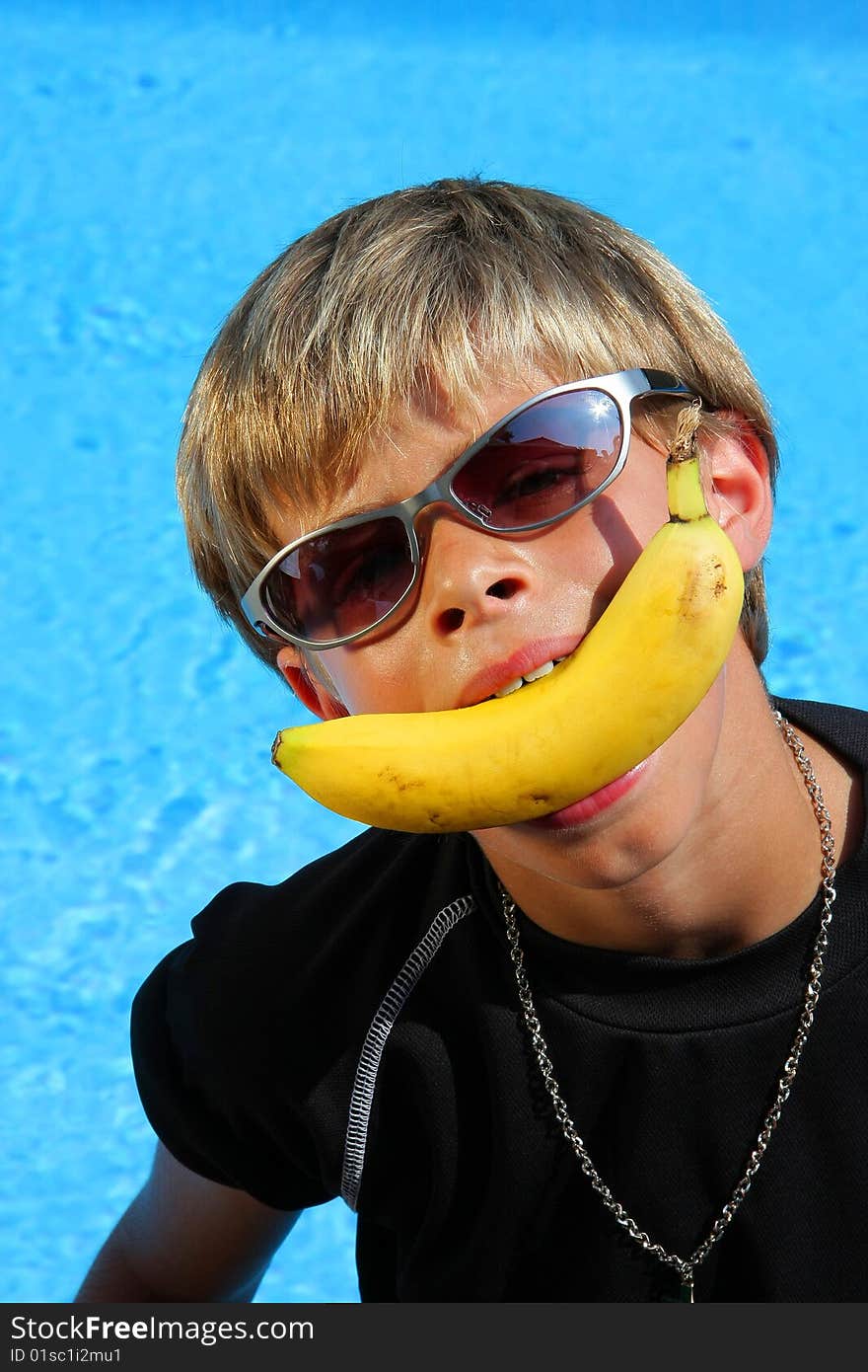 A 10 year old American - German boy with sun glasses sitting at a swimming pool in the summer sun joking with a banana between his teeth. A 10 year old American - German boy with sun glasses sitting at a swimming pool in the summer sun joking with a banana between his teeth