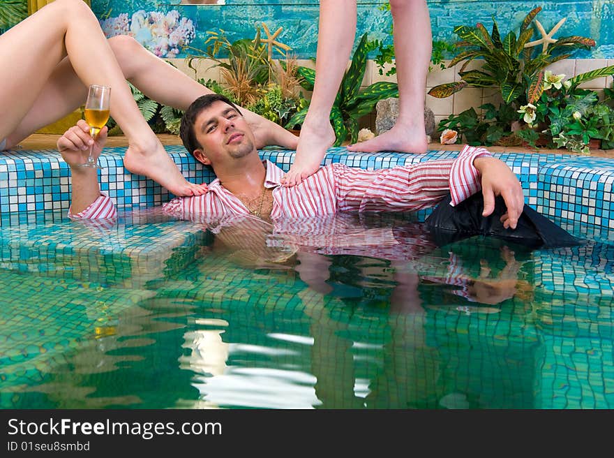 Young handsome man with a glass and the clothes in the pool surrounded by women's feet. Young handsome man with a glass and the clothes in the pool surrounded by women's feet