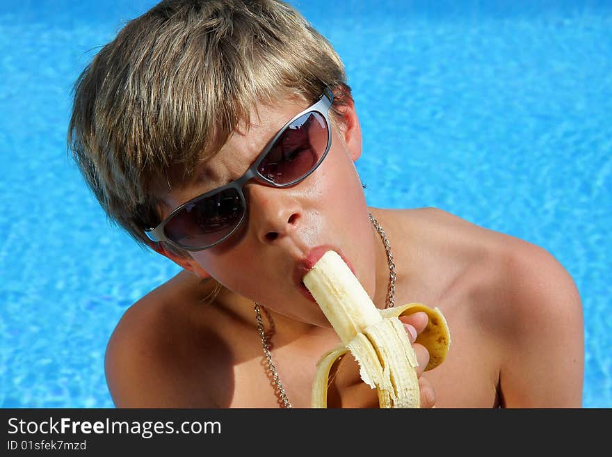 A 10 year old American - German boy with sun glasses sitting at a swimming pool in the summer sun eating a banana. A 10 year old American - German boy with sun glasses sitting at a swimming pool in the summer sun eating a banana