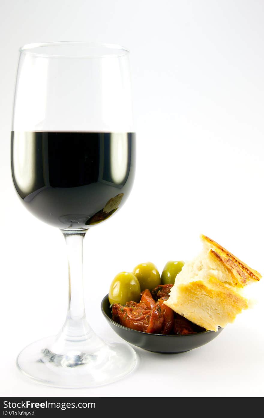 Pile of red sun dried tomatoes with three green olives and crusty bread in a small black dish with a glass of red wine on a plain background. Pile of red sun dried tomatoes with three green olives and crusty bread in a small black dish with a glass of red wine on a plain background
