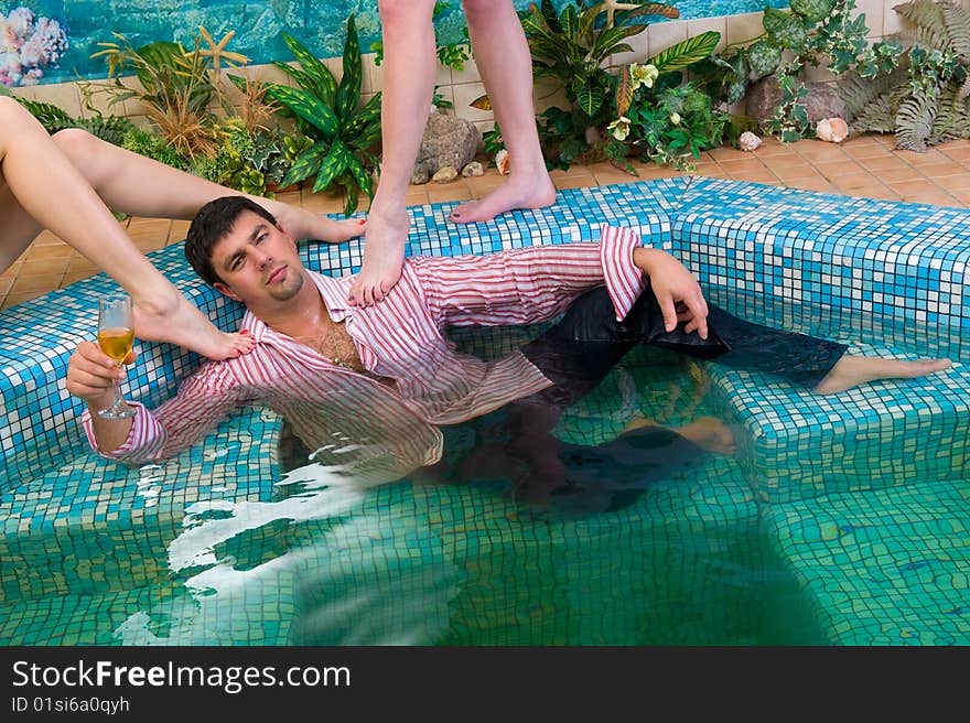 Young handsome man with a glass and the clothes in the pool surrounded by women's feet. Young handsome man with a glass and the clothes in the pool surrounded by women's feet