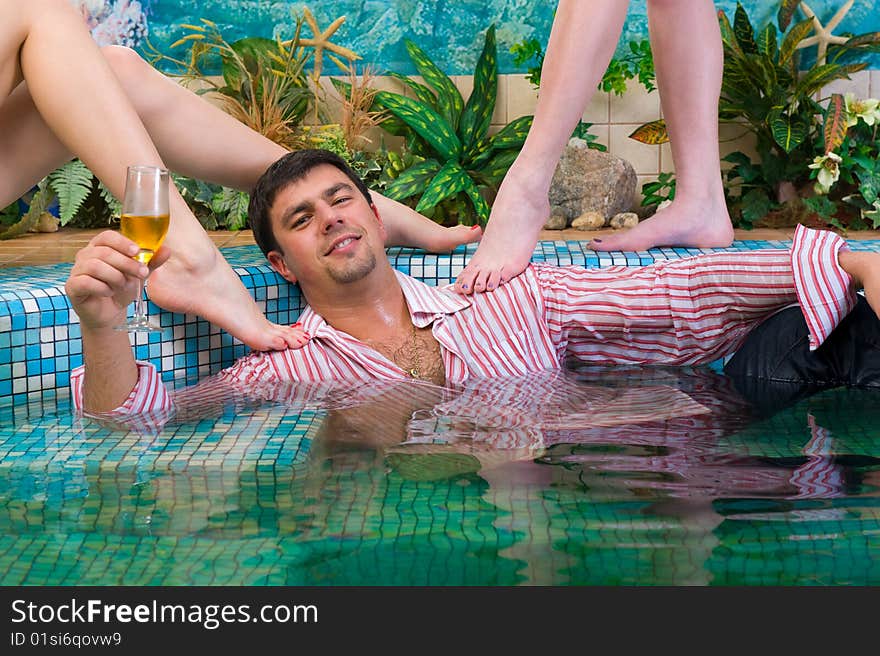 Young handsome man with a glass and the clothes in the pool surrounded by women's feet. Young handsome man with a glass and the clothes in the pool surrounded by women's feet