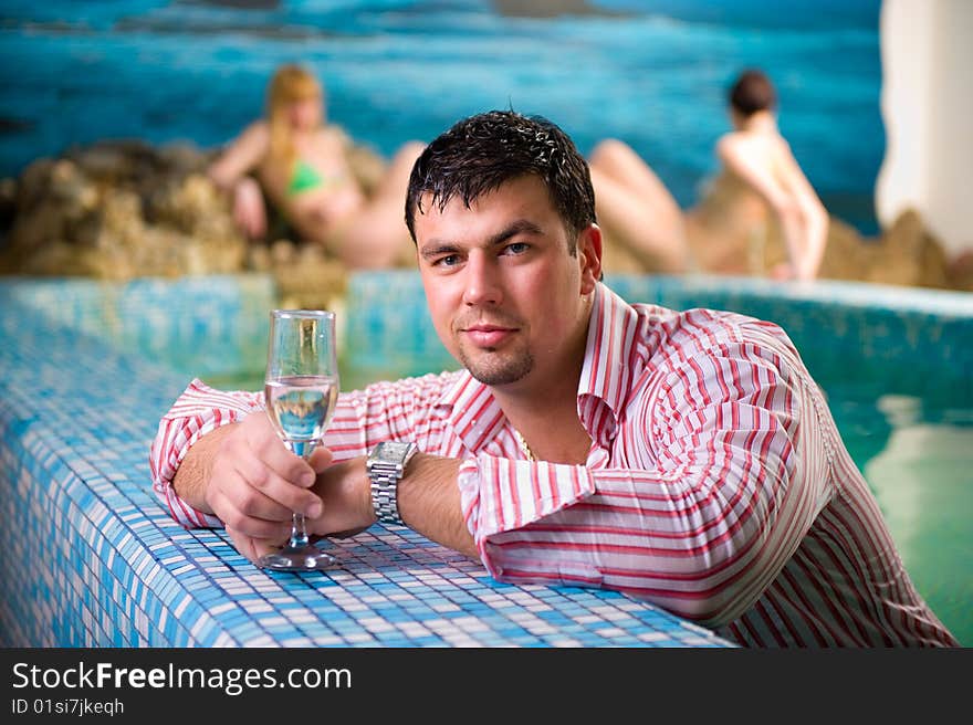 Portrait of a young man with a glass in the pool against the backdrop of two beautiful girls. Portrait of a young man with a glass in the pool against the backdrop of two beautiful girls