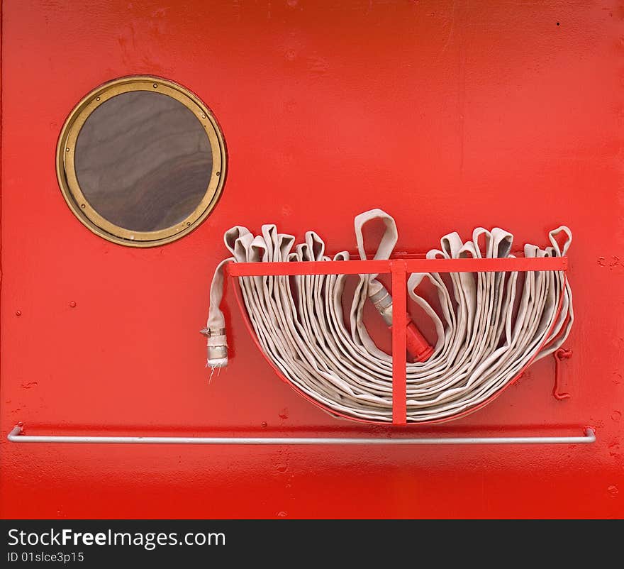 Closely cropped details on a red metal wall of a tugboat showing fire hose and round window. Closely cropped details on a red metal wall of a tugboat showing fire hose and round window