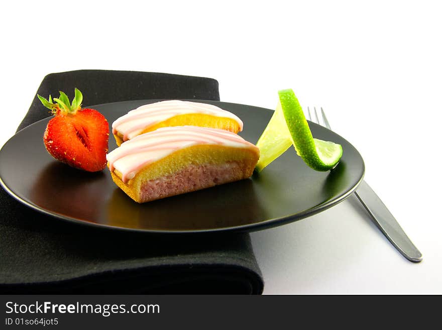 Two pink strawberry sliced cakes with icing on the top with a half strawberry and twist of lime on a black plate with a black napkin and fork on a white background. Two pink strawberry sliced cakes with icing on the top with a half strawberry and twist of lime on a black plate with a black napkin and fork on a white background
