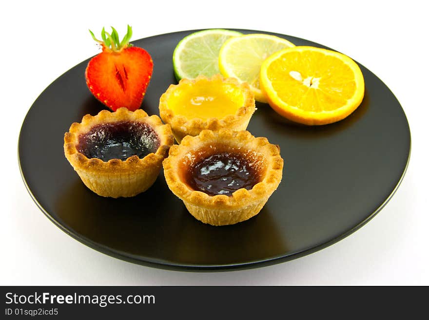 Red and yellow small jam tarts with slices of lemon, lime, orange and strawberry on a black plate on a white background. Red and yellow small jam tarts with slices of lemon, lime, orange and strawberry on a black plate on a white background