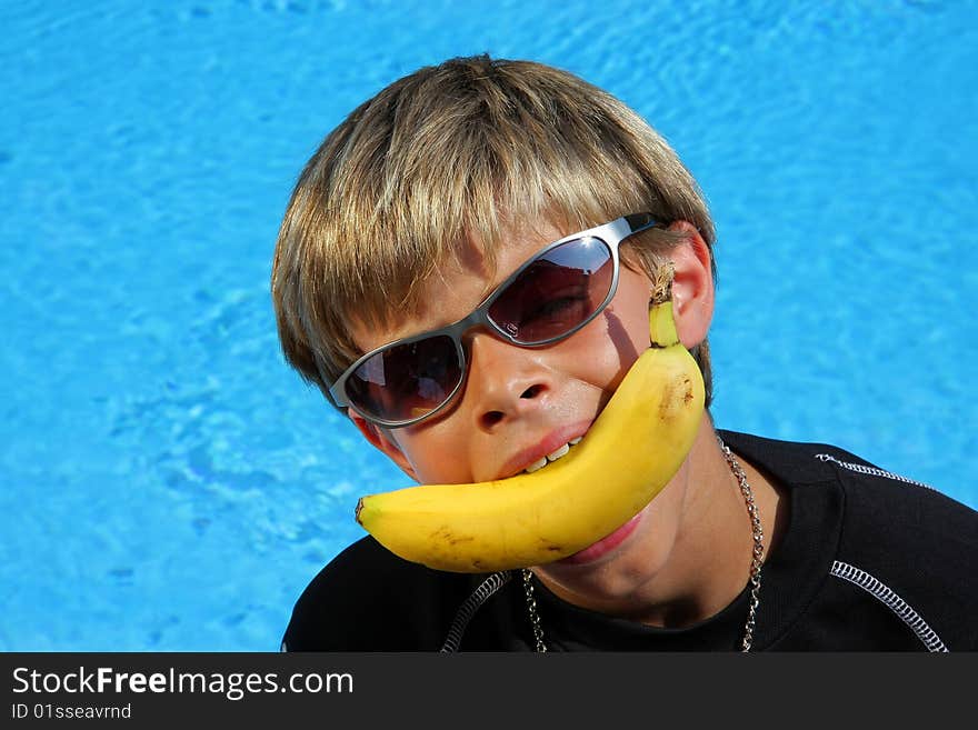 A 10 year old American - German boy with sun glasses joking with a banana in his mouth sitting at a swimming pool in the summer sun. A 10 year old American - German boy with sun glasses joking with a banana in his mouth sitting at a swimming pool in the summer sun