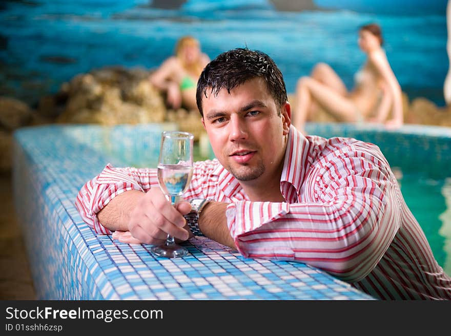 Portrait of a young man with a glass in the pool against the backdrop of two beautiful girls. Portrait of a young man with a glass in the pool against the backdrop of two beautiful girls